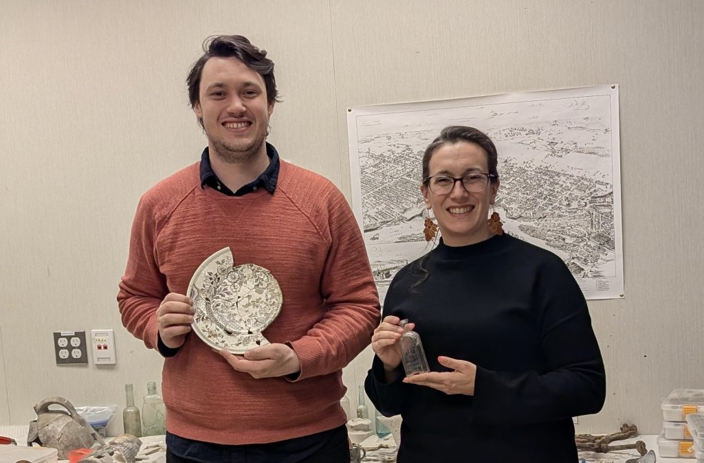 Job seeker Eamonn holding and artifact standing beside mentor Monica holding another artifact. They are standing in the archeology lab.